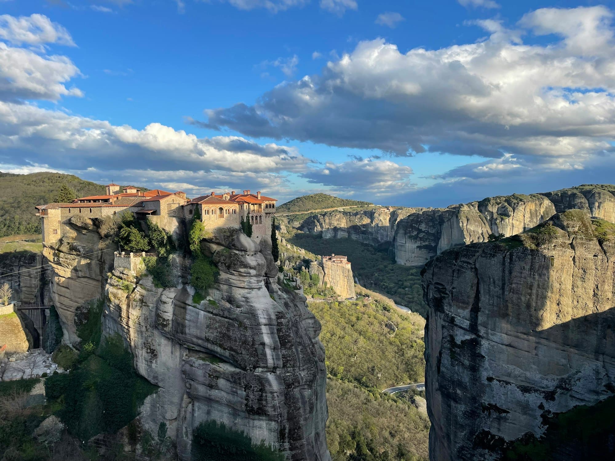 Meteora'da kumtaşı kayanın tepesinde kiremit çatılı manastır; arkada ikinci bir manastır ve kaya kuleleri.