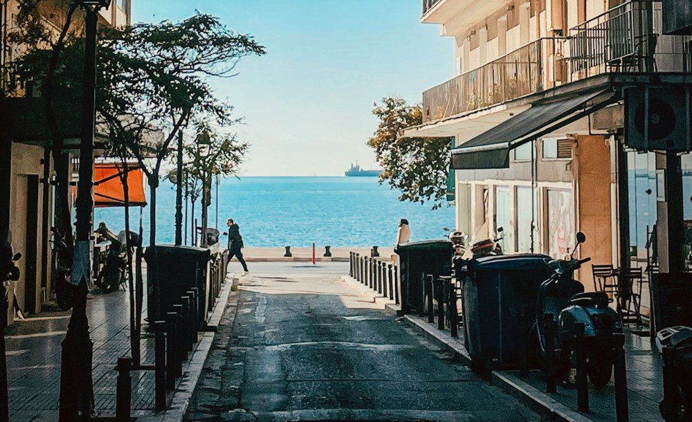 Στενό δρομάκι στο κέντρο της Θεσσαλονίκης με θέα προς τη θάλασσα και περαστικούς που περπατούν κάτω από το φως του ήλιου.